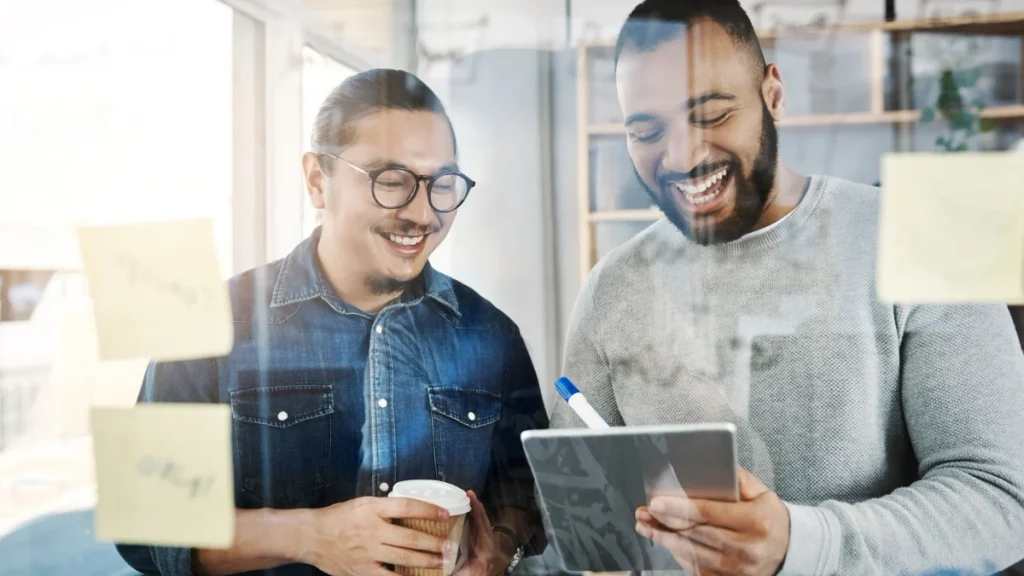 Two coworkers smile and review ideas on a tablet in a bright glass-walled office, with sticky notes on the glass and a coffee cup nearby.