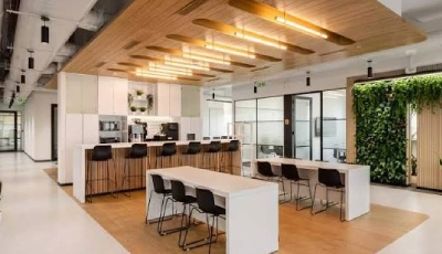 Modern office break area with a long bar counter, bar stools, and white tables for communal seating under warm wooden ceiling lights.